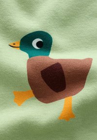 Kangas puuvillasta vaaleanvihreänä, jossa on ankkagraafinen kuva, teal-päillä, ruskealla vartalolla, keltaisella nokalla ja oransseilla jaloilla, kävelevässä asennossa.