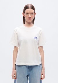 Λευκ ό βαμβακερό T-shirt με κοντά μανίκια, στρογγυλή λαιμόκοψη και μικρό μπλε λογότυπο στο αριστερό στήθος. Συνδυάζεται με ανοιχτό μπλε τζιν παντελόνι.