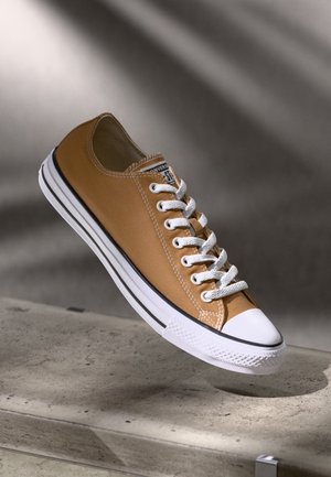 Brun lavtop lærreds-sneaker med hvide snørebånd og gummisål, svævende over en betonoverflade med blød skyggebaggrund.
