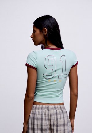 Frau mit langen dunklen Haaren, seitlich stehend, trägt ein hellblaues T-Shirt mit burgunderfarbenem Rand und der Nummer 91 auf dem Rücken sowie karierte Hose.