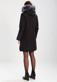 Parka negra de longitud hasta la rodilla con una capucha suave adornada con piel. Cuenta con mangas largas y un diseño recto, acompañada de botas negras hasta el tobillo.