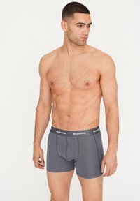 Calzoncillos tipo boxers para hombre en gris con un ajuste ceñido, featuring una cinturilla oscura con marca, diseño sin costuras y un compartimento contorneado para soporte.