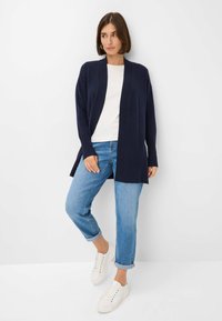 Cardigan lavorato a maglia blu con apertura frontale, polsini a costine e spacco laterale. Abbinato a jeans azzurri arrotolati e sneakers bianche.