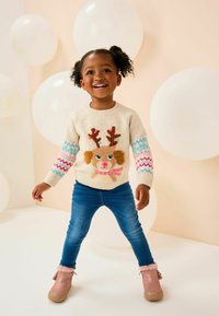 Cremefarbener Pullover mit einem Rentierdesign, bunten gemusterten Ärmeln, blauen Jeans und Boots mit rosa Fellbesatz. Der Hintergrund hat weiße Ballons.