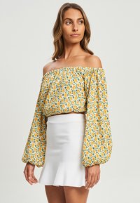 Top giallo scollato con motivo floreale e maniche lunghe a sbuffo, abbinato a una mini gonna bianca a campana.
