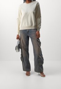 Persona vistiendo un sudadera crema con el texto "LOVE", jeans cargo desgastados, sandalias de tacón transparente y sosteniendo un pequeño bolso de mano plateado.