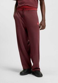 Maroon sweatpants with a contrasting red drawstring and waistband. Features side pockets and a small logo on the left leg. Smooth texture.