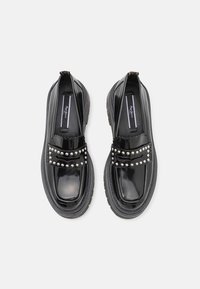 Paire de mocassins noirs brillants avec des semelles basses et épaisses et des clous argentés sur la sangle avant, vue de dessus sur fond blanc.