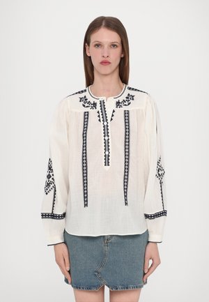 Blouse blanche à manches longues, avec des accents brodés noirs, fermeture à boutons et encolure arrondie. Associée à une jupe en denim.