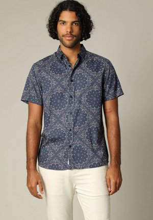 Chemise courte à manches en bleu marine, ornée de motifs floraux, avec un col boutonné et une texture de tissu lisse, associée à un pantalon de couleur claire.