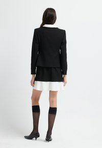 Kvinna står vänd bort, iklädd en svart kavaj, svart och vit lagerkjol, genomskinliga svarta knähöga strumpor och svarta låga klackskor.