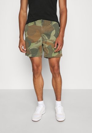 Uomo con pantaloncini mimetici verdi e marroni, t-shirt nera, calzini bianchi e sneakers bianche in piedi su sfondo bianco.