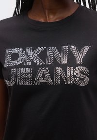 Svart t-skjorte med "DKNY JEANS" tekst i sølvfargede rundnagle, med klassisk rund hals og korte ermer, laget av mykt bomullsmateriale.