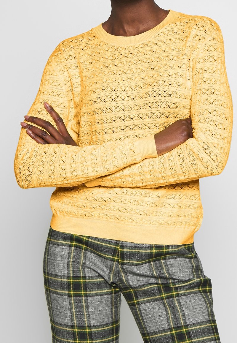 Personne portant un pull en maille jaune avec un motif en losanges et un pantalon à carreaux gris avec des rayures jaunes et vertes, les bras croisés.