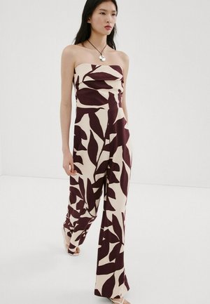 Mujer vistiendo un mono sin tirantes de pierna ancha con grandes patrones de hojas oscuras sobre un fondo claro, caminando contra un fondo blanco liso.