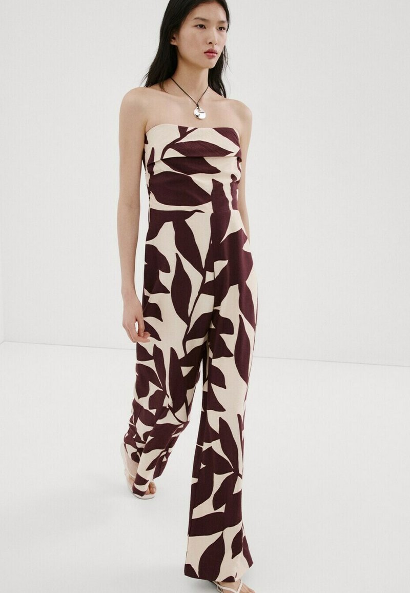 Žena v elegantnom overal s odhalenými ramenami a širokými nohavicami, zdobenom veľkými tmavými listovými vzormi na svetlom pozadí, kráča pred jednoduchým bielym pozadím.