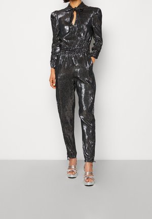 Femme portant une combinaison-robe noire brillante à manches longues avec un col noué en forme de nœud papillon et des sandales à talons hauts argentés, debout contre un mur blanc.