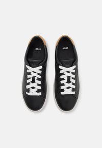 Sorte lærersneakers med hvite snørebånd, en beige lærerdetalj på hælen, og en hvit gummisåle. Logoet "BOSS" synlig på innsiden.