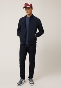 Giacca a zip blu navy sopra una t-shirt blu, jeans scuri e sneakers multicolori. Il cappello presenta un design bordeaux e bianco. Stile semplice e moderno.