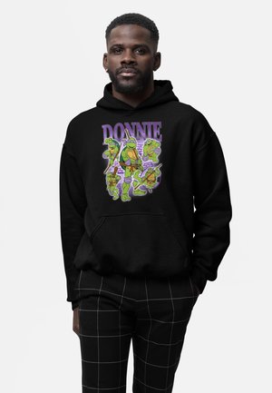 Homme portant un sweat à capuche noir avec cinq Tortues Ninja adolescents en dessin animé et le texte « DONNIE », assorti à un pantalon noir avec un motif quadrillé blanc.