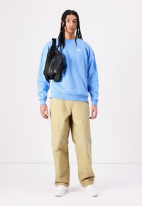 Kék pamut pulóver ribbelt mandzsettával, bézs nadrág texturált részletekkel, és egy fekete keresztbody táska. A modell egy semleges háttér előtt  áll.