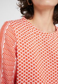Top de punto con un patrón geométrico en naranja y blanco, mangas cortas, escote redondo y una textura de tela suave.