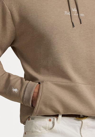 Felpa con cappuccio color sabbia dallo stesso morbido, caratterizzata da un piccolo logo bianco sulla manica e da lacci. Abbinata a pantaloni chiari.