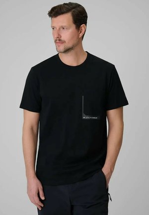 Mężczyzna w czarnym krótkim T-shircie z małą grafiką na kieszeni i ciemnych spodniach, stojący na jednolitym szarym tle.