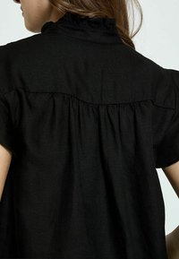 Čierny top s volánovým golierom, krátkymi rukávmi a jemnou textúrovanou tkaninou. Zadná strana má zjednodušené šitie s naberaním pre zvýšený objem.