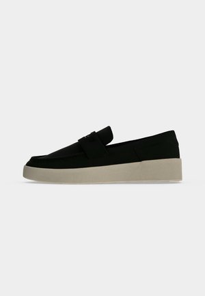 Μαύρο παπούτσι τύπου slip-on με χοντρή ανοιχτόμπεζ σόλα, χαμηλό τακούνι και λεπτομέρεια με λουράκι πάνω στο επάνω μέρος, σε απλό ανοιχτό γκρι φόντο.