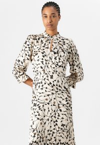 Langärmliges Kleid in Creme mit einem floralen Muster in Schwarz und Braun, hohem Kragen mit Schlüssellochöffnung und gerafften Ärmeln.