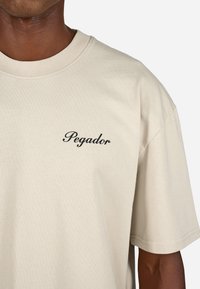 Beżowa bawełniana koszulka z krótkim rękawem i okrągłym dekoltem. Na lewej piersi znajduje się czarny napis "Pegador" w kursywie.