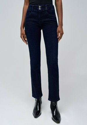 Dunkelblaue Jeans aus Denim mit geradem Bein, ausgestattet mit zwei frontalen Taschen und zwei Knopfverschlüssen in der Taille. Getragen mit schwarzen Stiefeletten.