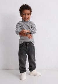 Un tout-petit souriant avec des cheveux bouclés, ajustant sa manche, portant un col roulé rayé noir et blanc, un jean noir et des baskets blanches, se tenant devant un mur blanc.