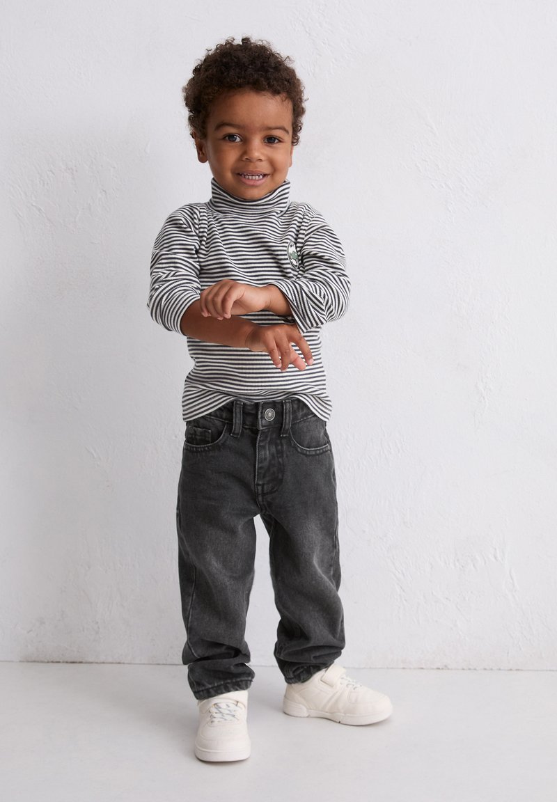 Un tout-petit souriant avec des cheveux bouclés, ajustant sa manche, portant un col roulé rayé noir et blanc, un jean noir et des baskets blanches, se tenant devant un mur blanc.