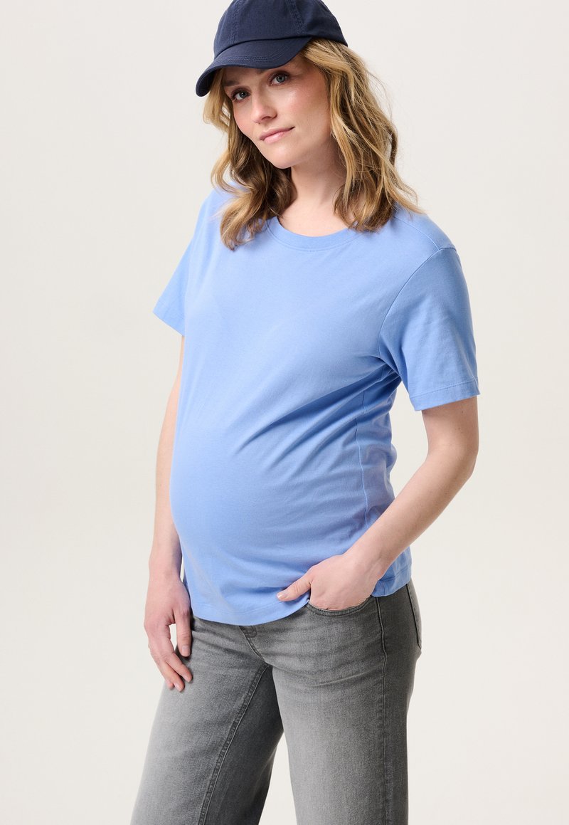 Lichtblauw katoenen zwangerschaps T-shirt met korte mouwen, ronde hals en een relaxte pasvorm, gecombineerd met grijze jeans en een marineblauwe pet.