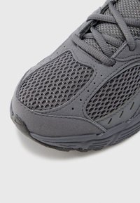 Γκρι αθλητικό παπούτσι με υλικά από δίχτυ και σουέτ, υφή στην επάνω επιφάνεια, και επενδυμένη γλώσσα με κλείσιμο με κορδόνια. Σχεδιασμένο για απόδοση.