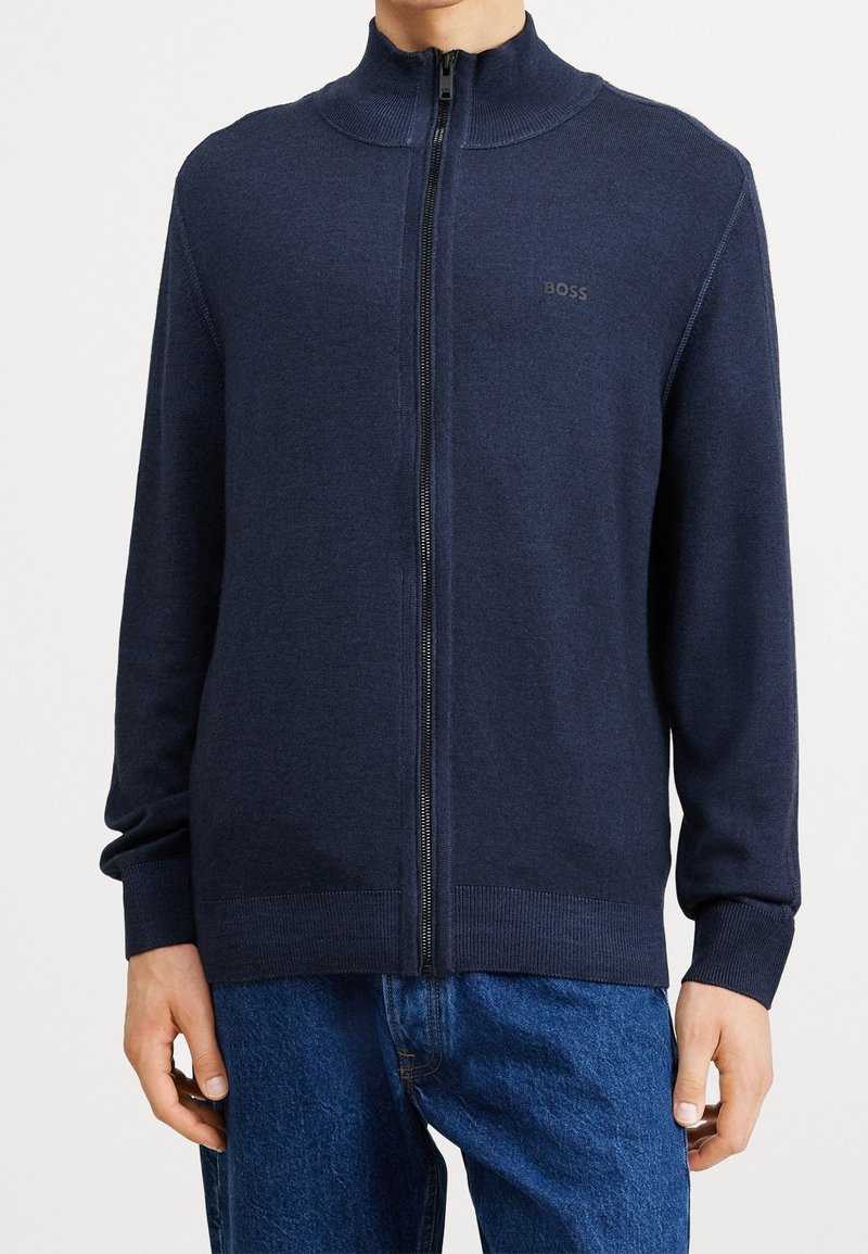 Man som bär en navyblå zip-up kofta med ribbad krage, muddar och fåll, i kombination med blå denimjeans.
