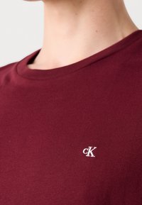 Maročná bavlněná trička s kulatým výstřihem a malým bílým vyšitým logem ‚cK‘ na levé hrudi. Hladká struktura, jednoduchý design.