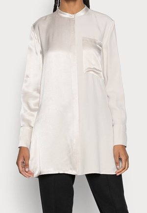 Blouse tunique en satin à manches longues avec col mandarin et poche avant, portée sur un pantalon noir, visible des épaules aux cuisses.