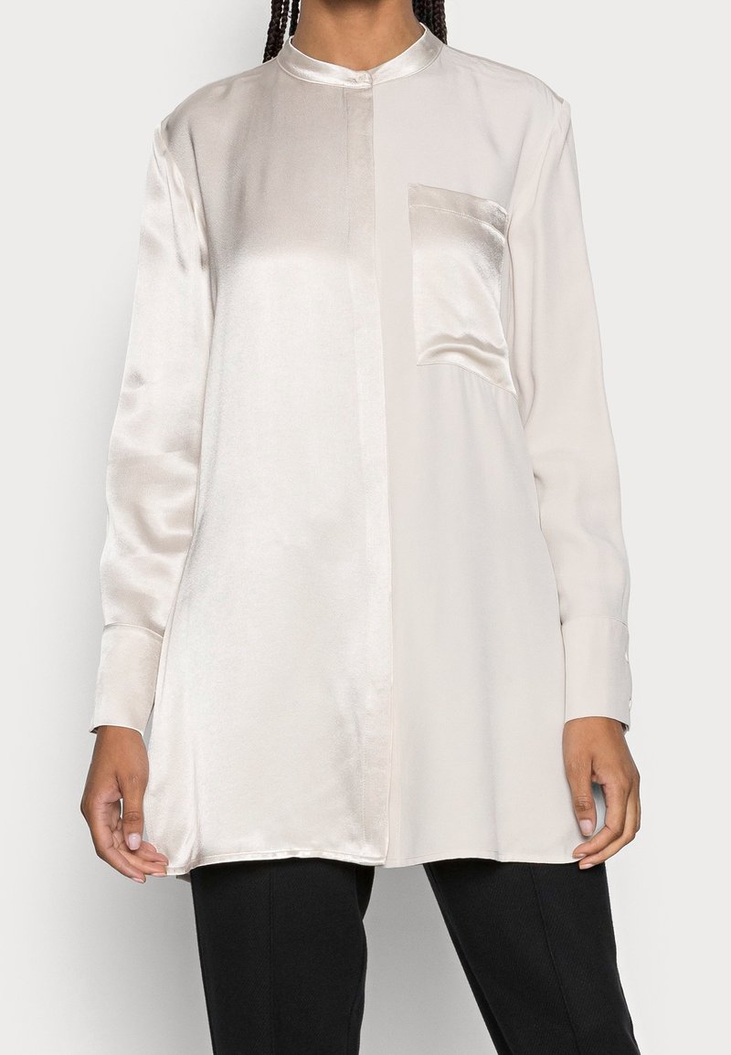 Blouse tunique en satin à manches longues avec col mandarin et poche avant, portée sur un pantalon noir, visible des épaules aux cuisses.