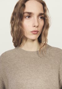 Maglione in lana beige con scollo rotondo e polsini a coste, caratterizzato da una texture morbida e una finitura liscia, indossato da una modella su uno sfondo neutro.