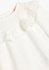 Weißes Langarmoberteil mit rüschem Ausschnitt und filigranen Spitzenverzierungen entlang der Rüsche, aus weichem Textilmaterial gefertigt.