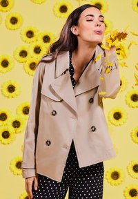 Beige trench coat with double-breasted design, large buttons, and wide collar. Model in polka dot pants, with yellow petals in a floral background.