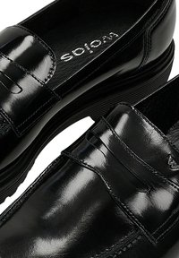 Mocassini penny in pelle nera lucida con suole spesse e punte arrotondate, caratterizzati da cuciture visibili e un piccolo logo metallico.