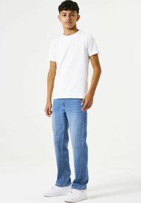 Camiseta corta blanca de manga corta, vaqueros rectos de color azul claro y zapatillas blancas. El modelo está de pie contra un fondo blanco liso.