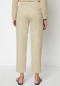 Pantalon beige à jambes larges avec une texture lisse et une coupe ajustée. Comprend une poche arrière et des ourlets avec revers, porté avec des tongs noires.