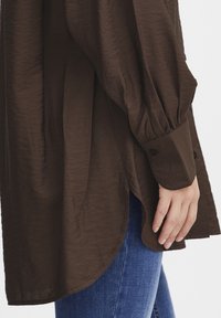 Brown, long-sleeve blouse with a textured finish, featuring a curved hem and buttoned cuffs. Paired with blue denim jeans.
