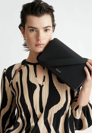Pochette noire texturée avec une bandoulière diagonale, tenue contre un modèle portant une blouse beige et noire à motifs avec des plis et des découpes.
