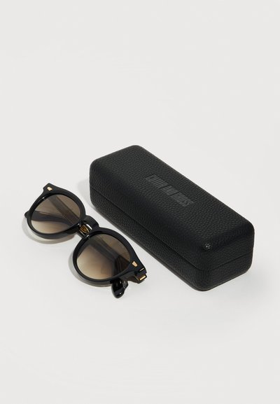 Μαύρα στρογγυλά γυαλιά ηλίου με σκούρους φακούς δίπλα σε μαύρη, υφασμάτινη ορθογώνια θήκη σε λευκό φόντο.